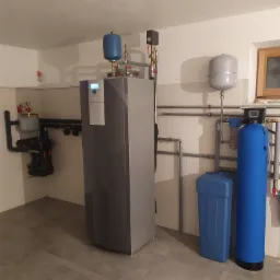 Szara instalacja grzewcza z dwoma niebieskimi zbiornikami, czarnymi rurami w izolacji oraz filtrem wody w niebieskiej obudowie, zamontowana w kotłowni z beżowymi płytkami na ścianach i szarą podłogą.