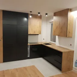 Nowoczesna kuchnia z czarnymi szafkami dolnymi i drewnopodobnymi górnymi, punktowe oświetlenie sufitowe, jasna podłoga, widok na okno.