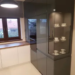 Nowoczesna kuchnia z szarymi, lakierowanymi szafkami, przeszkloną witryną z filiżankami i białymi dolnymi szafkami, oświetlona wiszącą lampą.