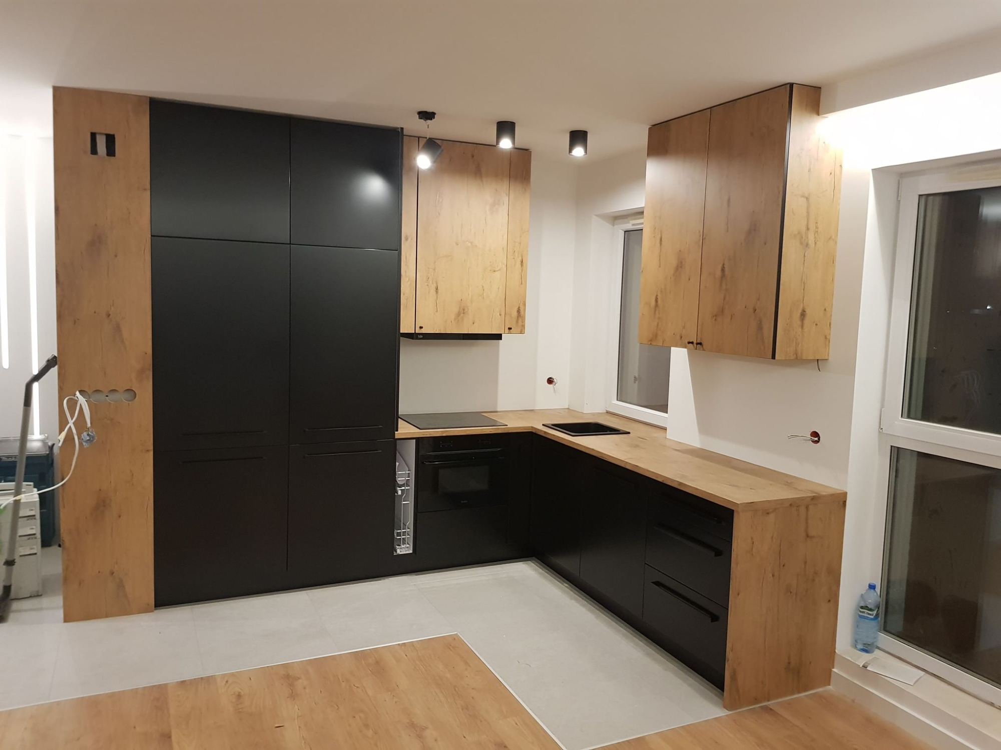 Nowoczesna kuchnia z czarnymi szafkami dolnymi i drewnopodobnymi górnymi, punktowe oświetlenie sufitowe, jasna podłoga, widok na okno.