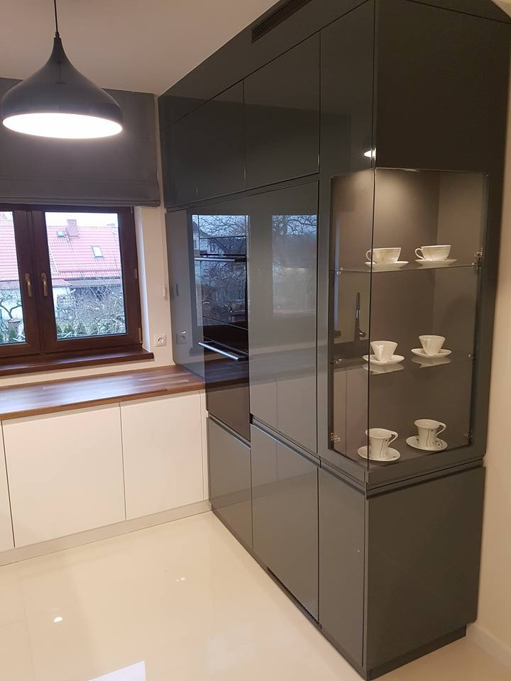 Nowoczesna kuchnia z szarymi, lakierowanymi szafkami, przeszkloną witryną z filiżankami i białymi dolnymi szafkami, oświetlona wiszącą lampą.