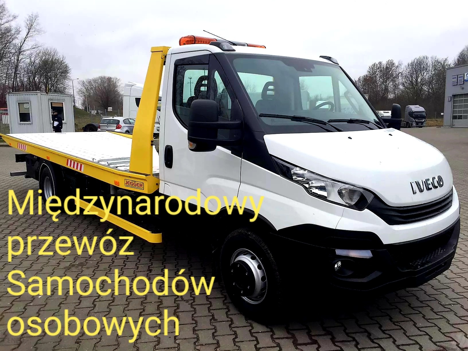 Biała laweta marki Iveco z żółtym podnośnikiem, zaparkowana na brukowanym placu przed budynkiem firmy transportowej.