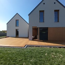 FREEDOM HANNA MALAWSKA - Rozległy taras z desek egzotycznych przylegający do nowoczesnego domu z szarą elewacją, widoczne drobne kamienie oddzielające taras od trawnika, jasne niebo.