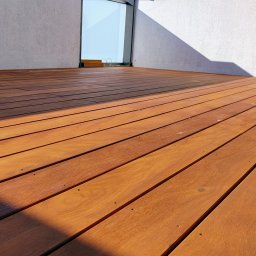 FREEDOM HANNA MALAWSKA - Nowoczesny taras z egzotycznego drewna, deski ułożone równolegle, widoczne mocowania. Ściana w tle z oknem i padającym cieniem. Ciepłe oświetlenie podkreśla strukturę drewna.