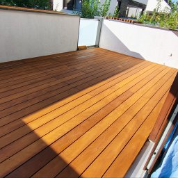 FREEDOM HANNA MALAWSKA - Jasnobrązowy taras z desek, widok z góry, z fragmentem okna balkonowego po prawej. Ściany balkonu białe, w tle zieleń i zabudowa.