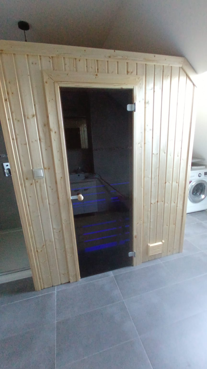 Wnętrze sauny fińskiej z drewnianą obudową i szklanymi drzwiami, widok z zewnątrz na podświetlone na niebiesko ławki, w tle pralka i łazienka.