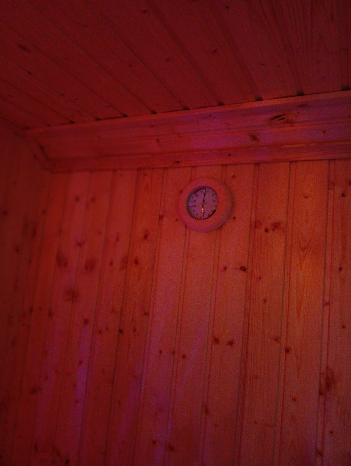 Wnętrze sauny wyłożone jasnym drewnem, oświetlone czerwonym światłem, z okrągłym termometrem/higrometrem zamontowanym na ścianie.