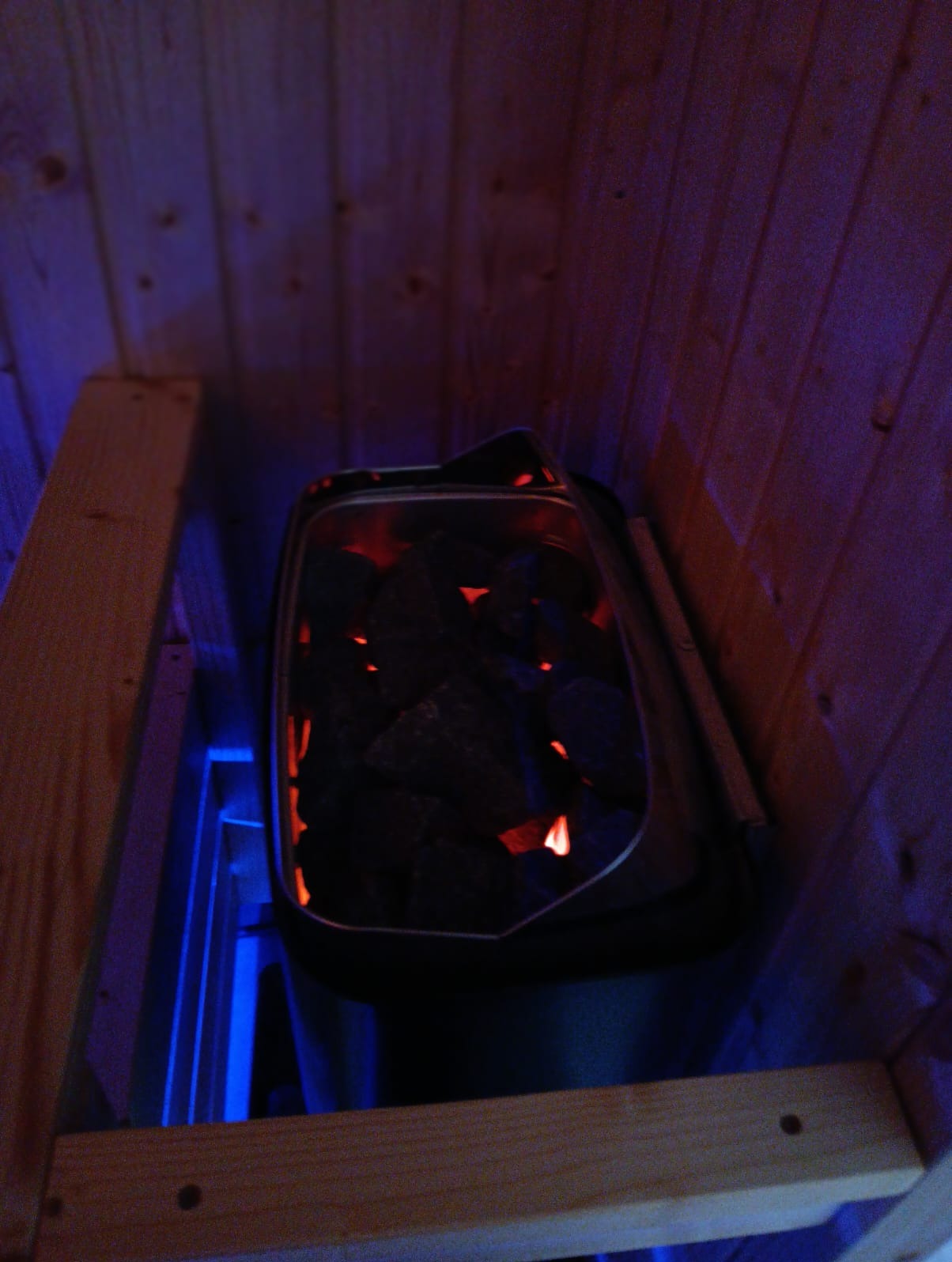 Wnętrze sauny z piecem wypełnionym rozgrzanymi kamieniami, oświetlonymi od dołu niebieskim światłem, widoczne drewniane deski boazerii i fragment konstrukcji ławki.