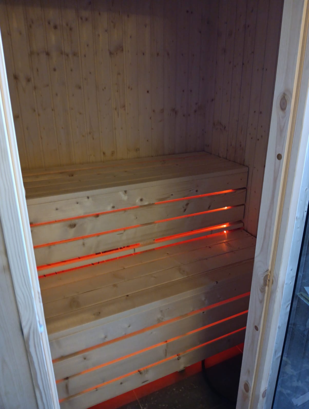 Wnętrze sauny fińskiej z drewnianą boazerią i ławami, podświetlonymi pomarańczowym światłem, widoczne przez otwarte drzwi.