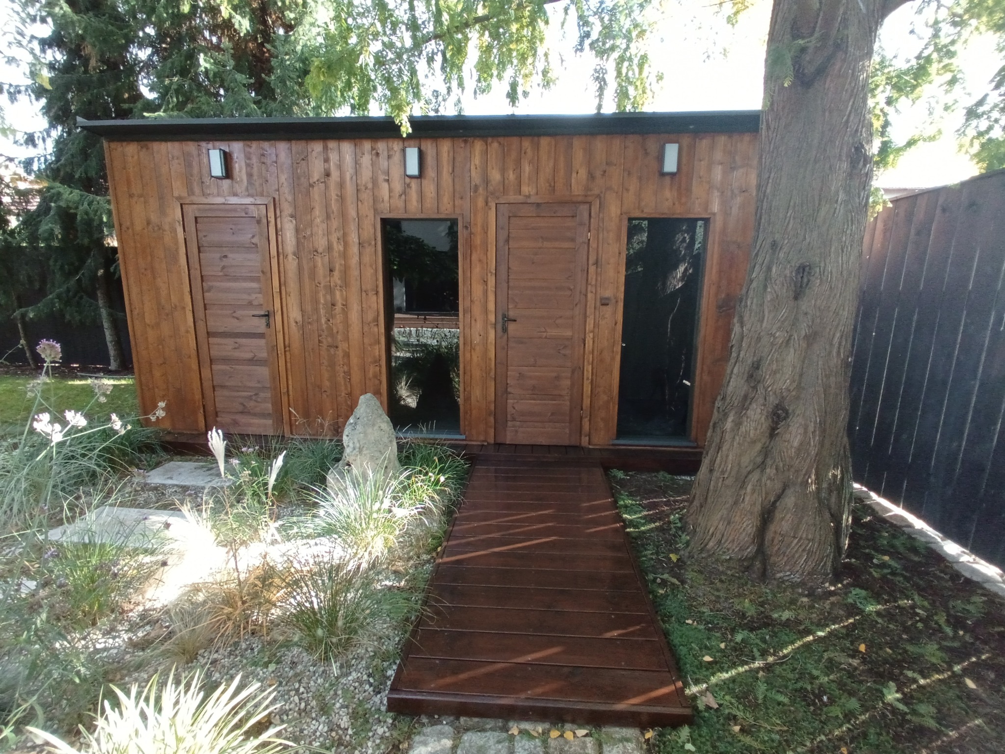 Drewniana sauna z dwoma wejściami i oknami, otoczona zielenią i drzewem. Do sauny prowadzi drewniany chodnik. Widoczne oświetlenie zewnętrzne.