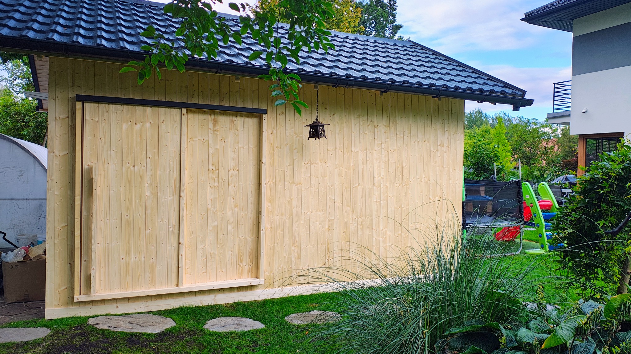 Drewniany domek warsztatowy z przesuwnymi drzwiami i czarnym dachem, oświetlony wiszącą latarenką, otoczony zielenią i kamienną ścieżką w ogrodzie.