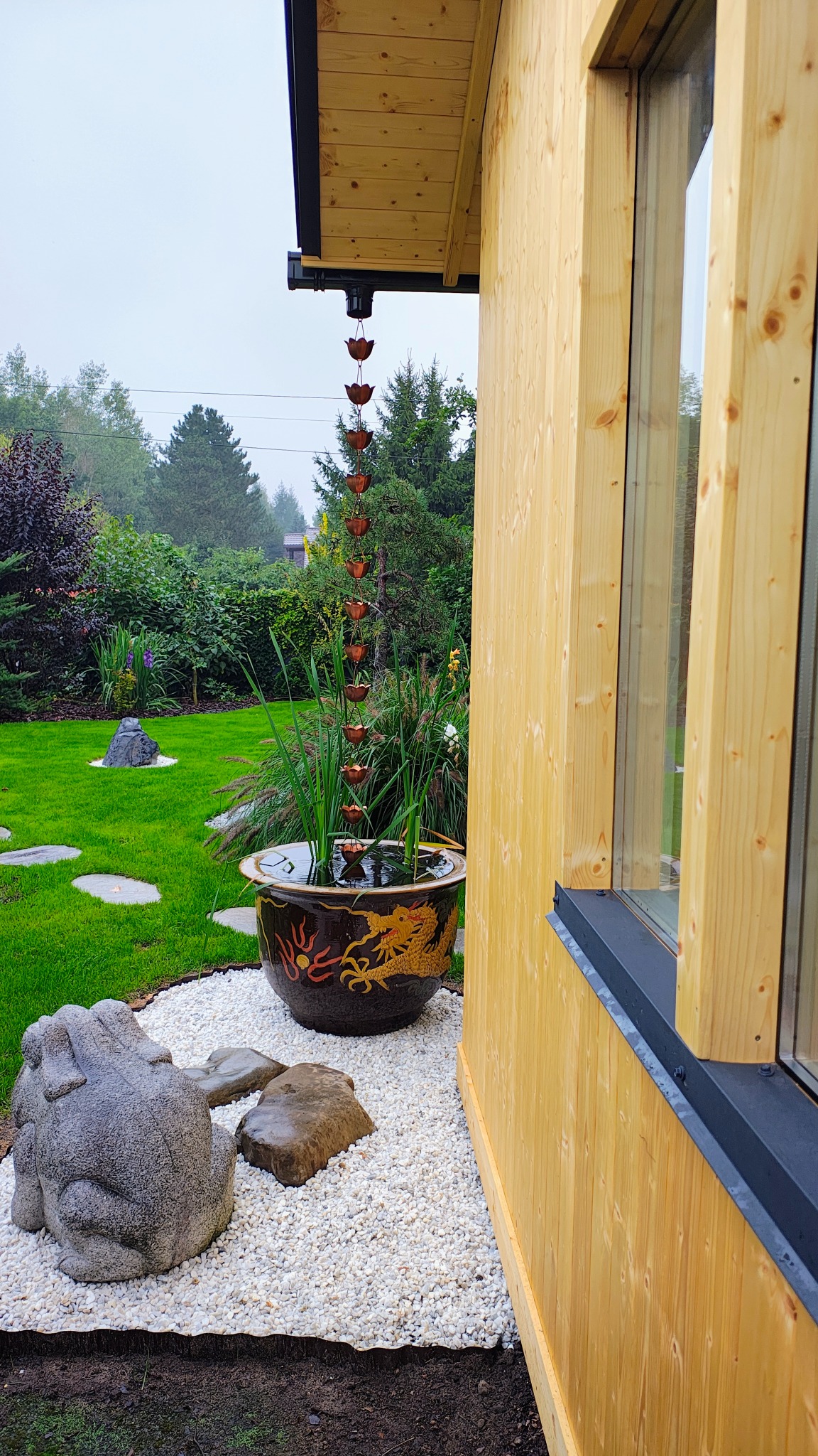 Drewniany domek z oknem i łańcuchem deszczowym nad ozdobną donicą z motywem smoka, otoczony białym żwirem i zielenią ogrodu.