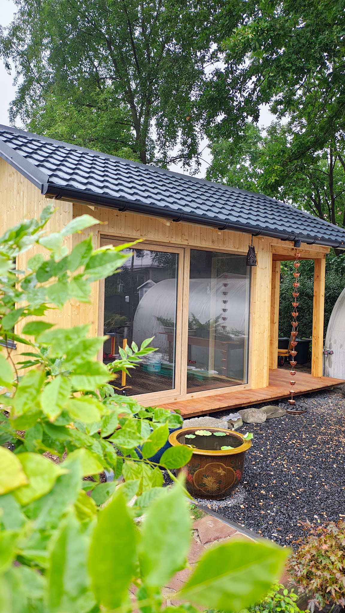 Nowoczesny domek warsztatowy z drewna, z dużymi oknami i czarną dachówką, otoczony zielenią i ozdobnym oczkiem wodnym. Widoczna dekoracyjna rynna łańcuchowa.