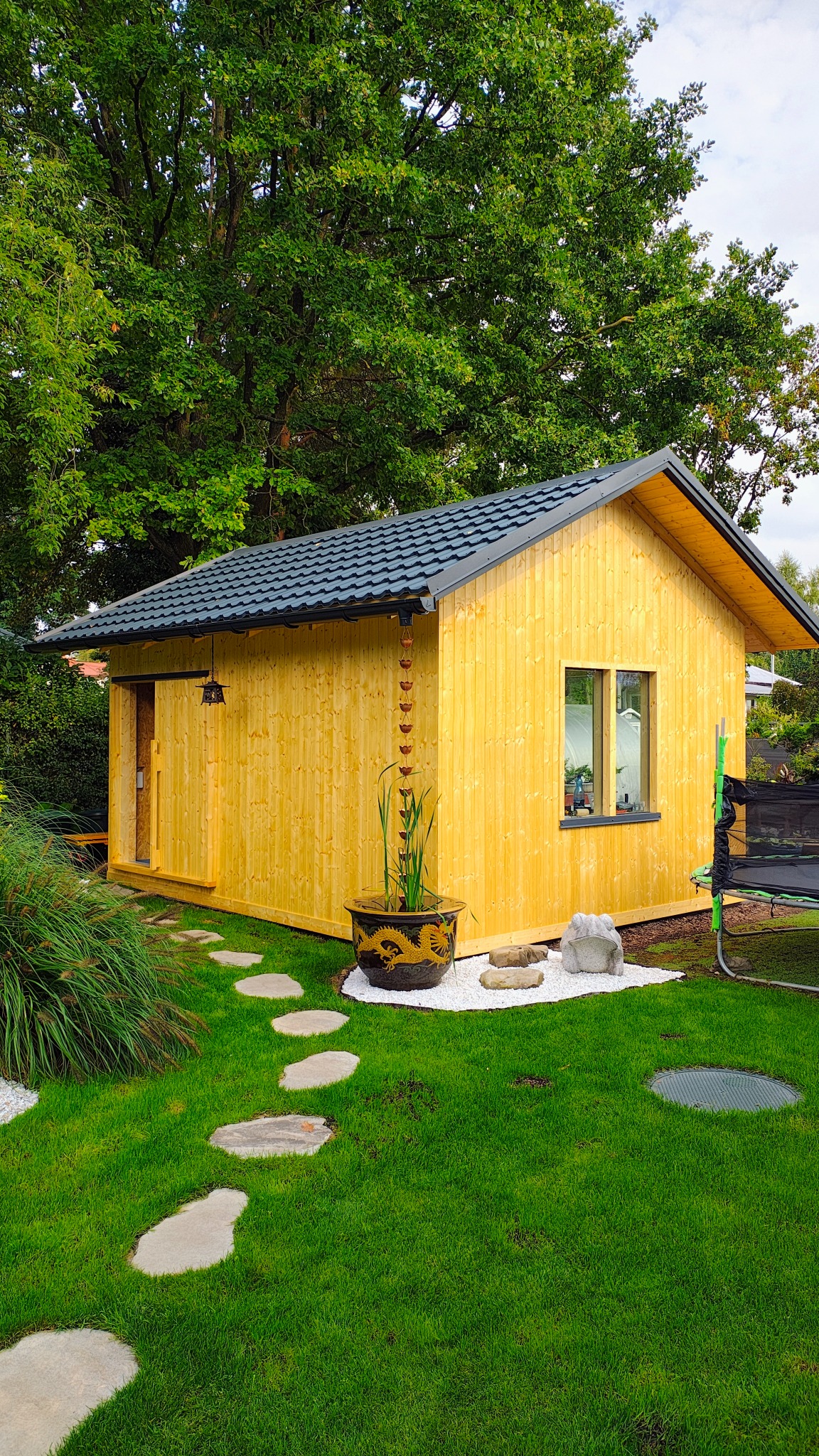 Żółty domek warsztatowy z szarą dachówką, widoczny z perspektywy ogrodu z kamienną ścieżką. Dekoracyjny łańcuch deszczowy i donica z motywem smoka dodają unikalności.