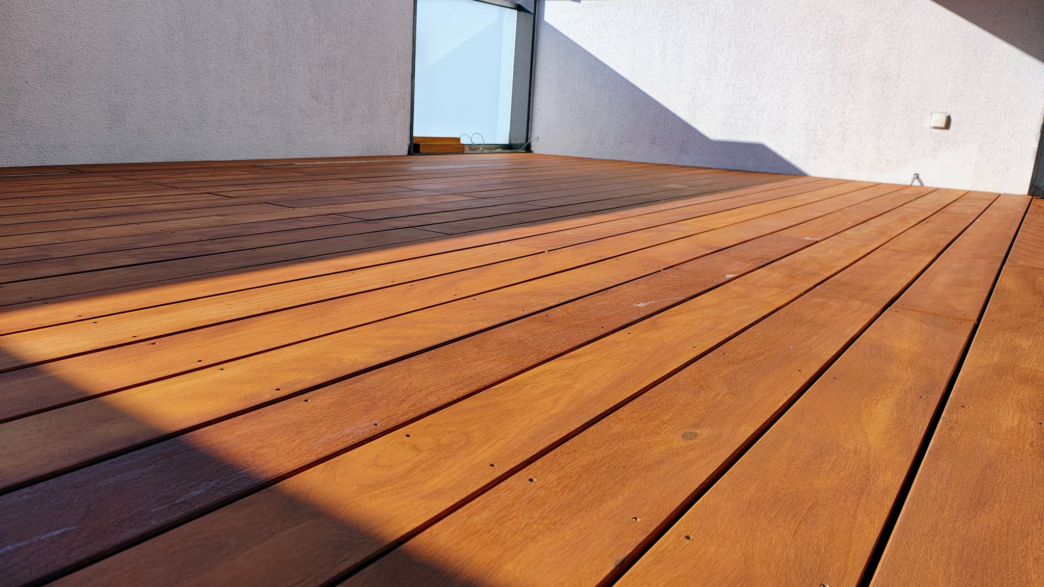 Nowoczesny taras z egzotycznego drewna, deski ułożone równolegle, widoczne mocowania. Ściana w tle z oknem i padającym cieniem. Ciepłe oświetlenie podkreśla strukturę drewna.
