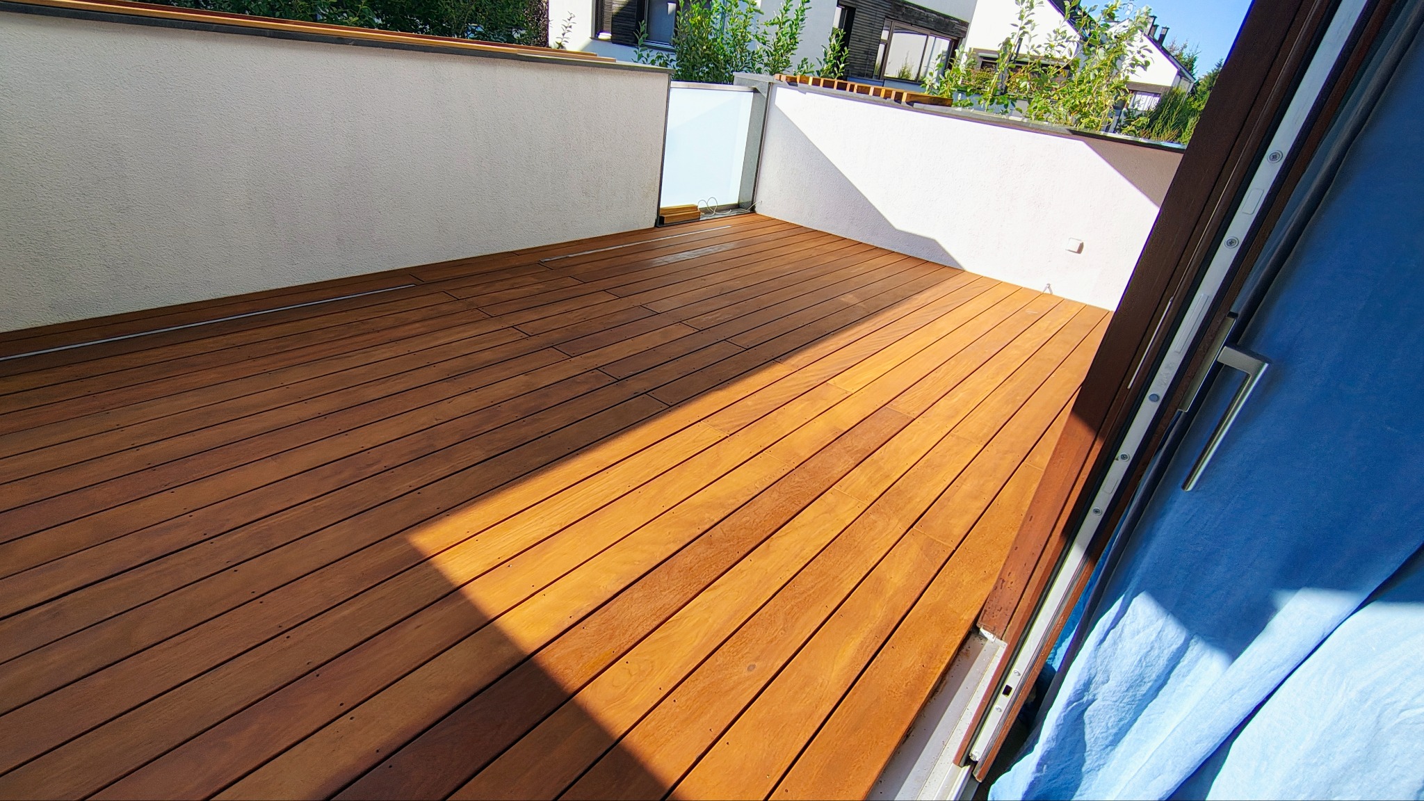 Jasnobrązowy taras z desek, widok z góry, z fragmentem okna balkonowego po prawej. Ściany balkonu białe, w tle zieleń i zabudowa.