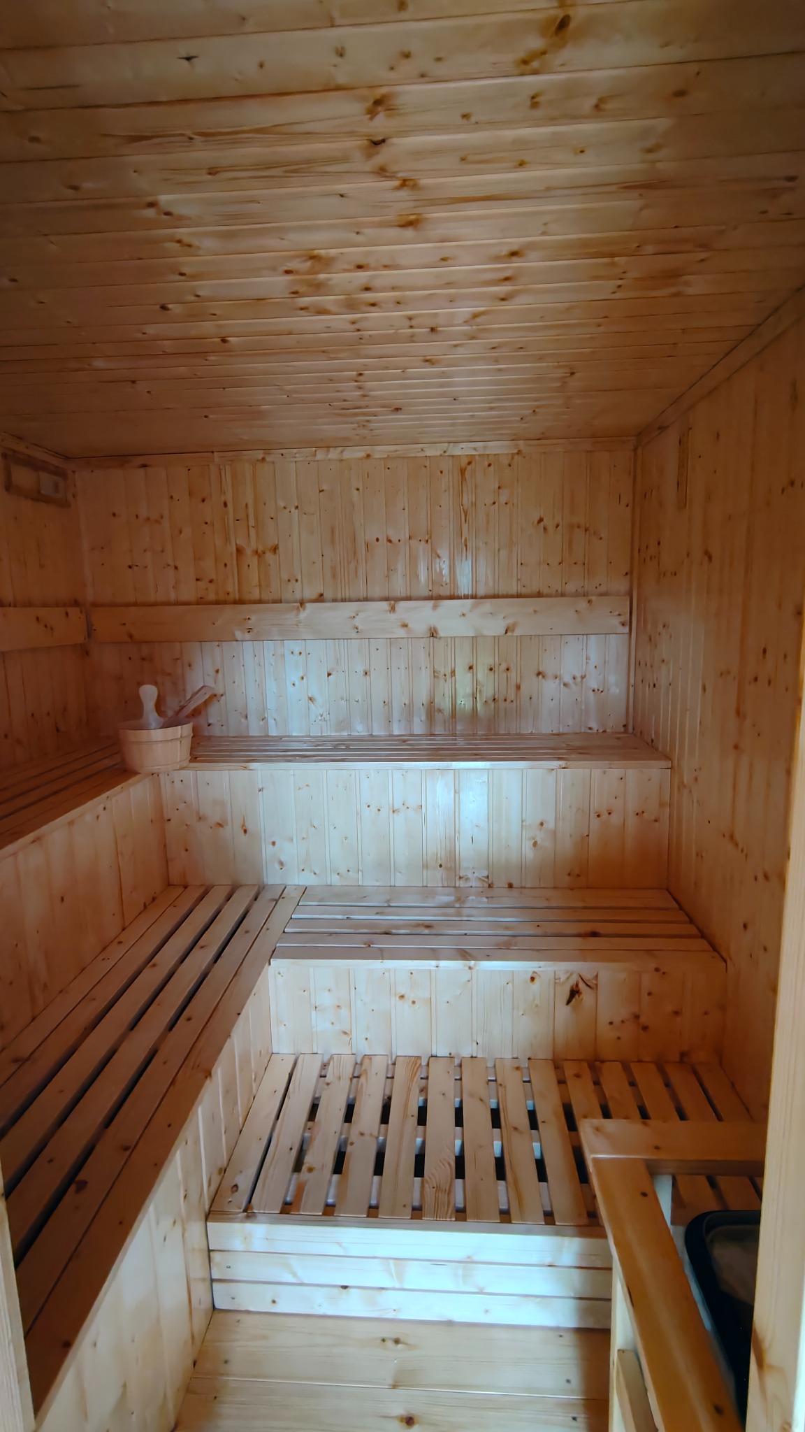 Wnętrze sauny z jasnego drewna: ławy schodkowe, drewniane ściany i sufit. Na górnej ławie wiaderko z chochlą. Widok z perspektywy wejścia.
