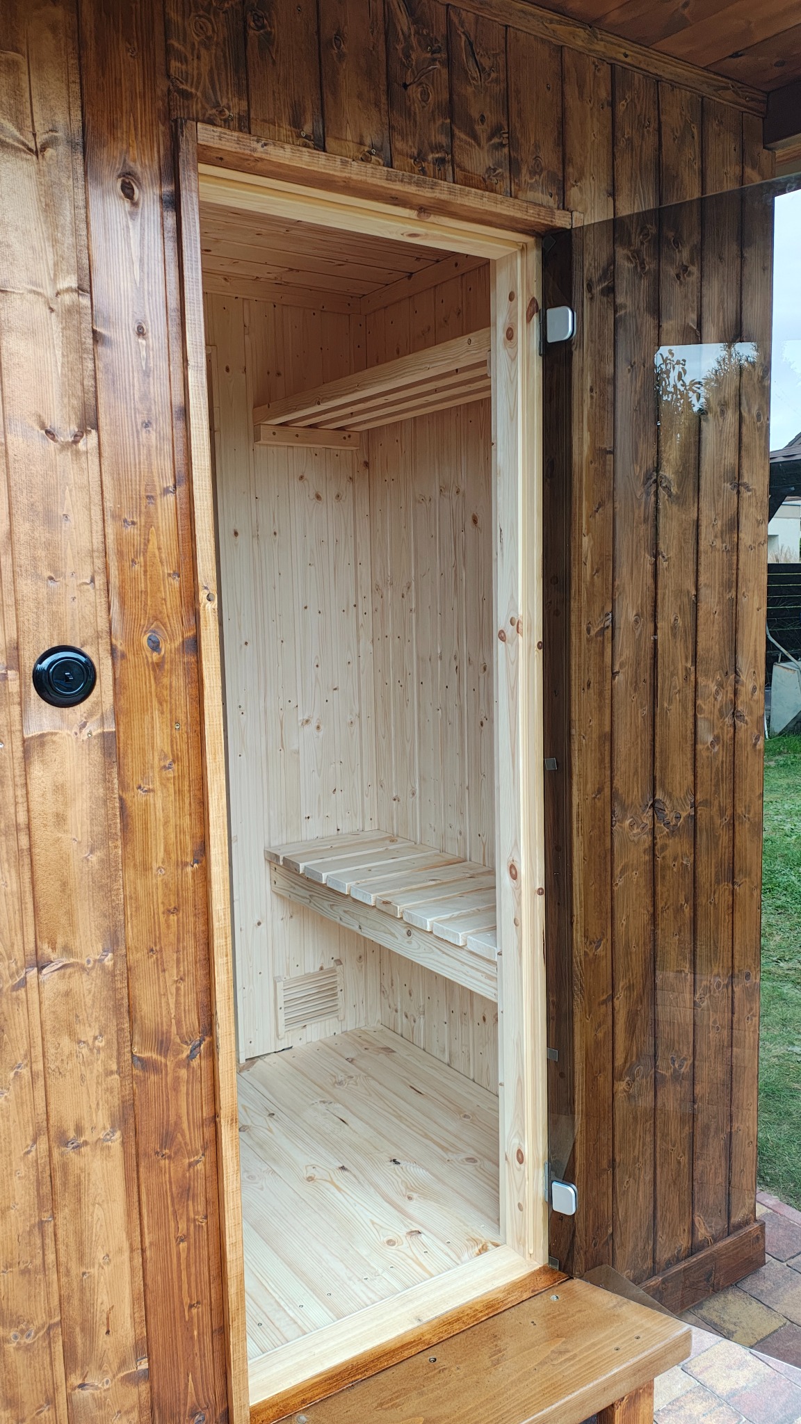 Otwarta, drewniana sauna z przedsionkiem. Widoczne jasne, drewniane wnętrze z ławkami i ciemniejsze, lakierowane deski na zewnątrz. Drzwi częściowo przeszklone.