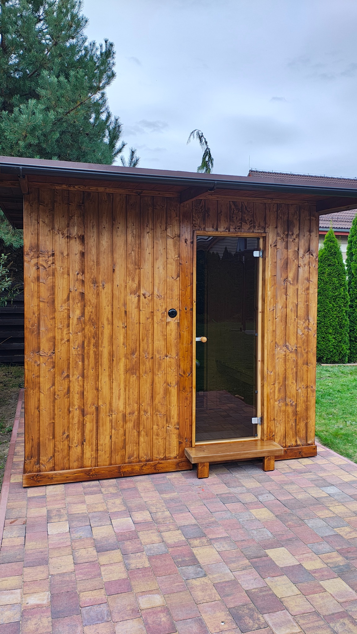 Drewniana sauna ogrodowa z przeszklonymi drzwiami, widoczna z zewnątrz na tle zieleni i kostki brukowej. Charakterystyczne pionowe deski i ciemne wykończenia.
