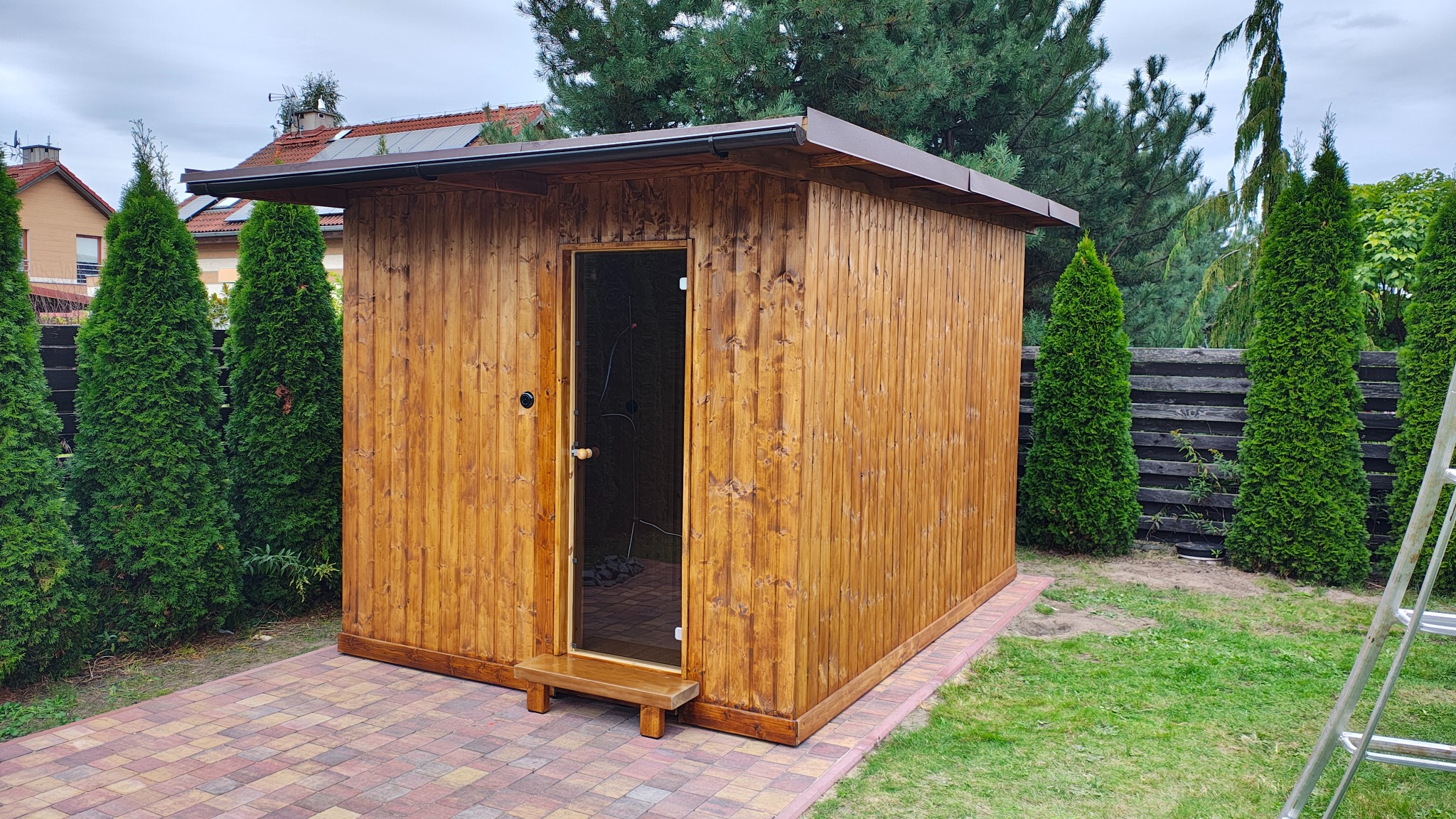 Drewniana sauna z przedsionkiem, z otwartymi drzwiami, na kostce brukowej, otoczona zielenią. Widoczna drabina oparta o ogrodzenie.