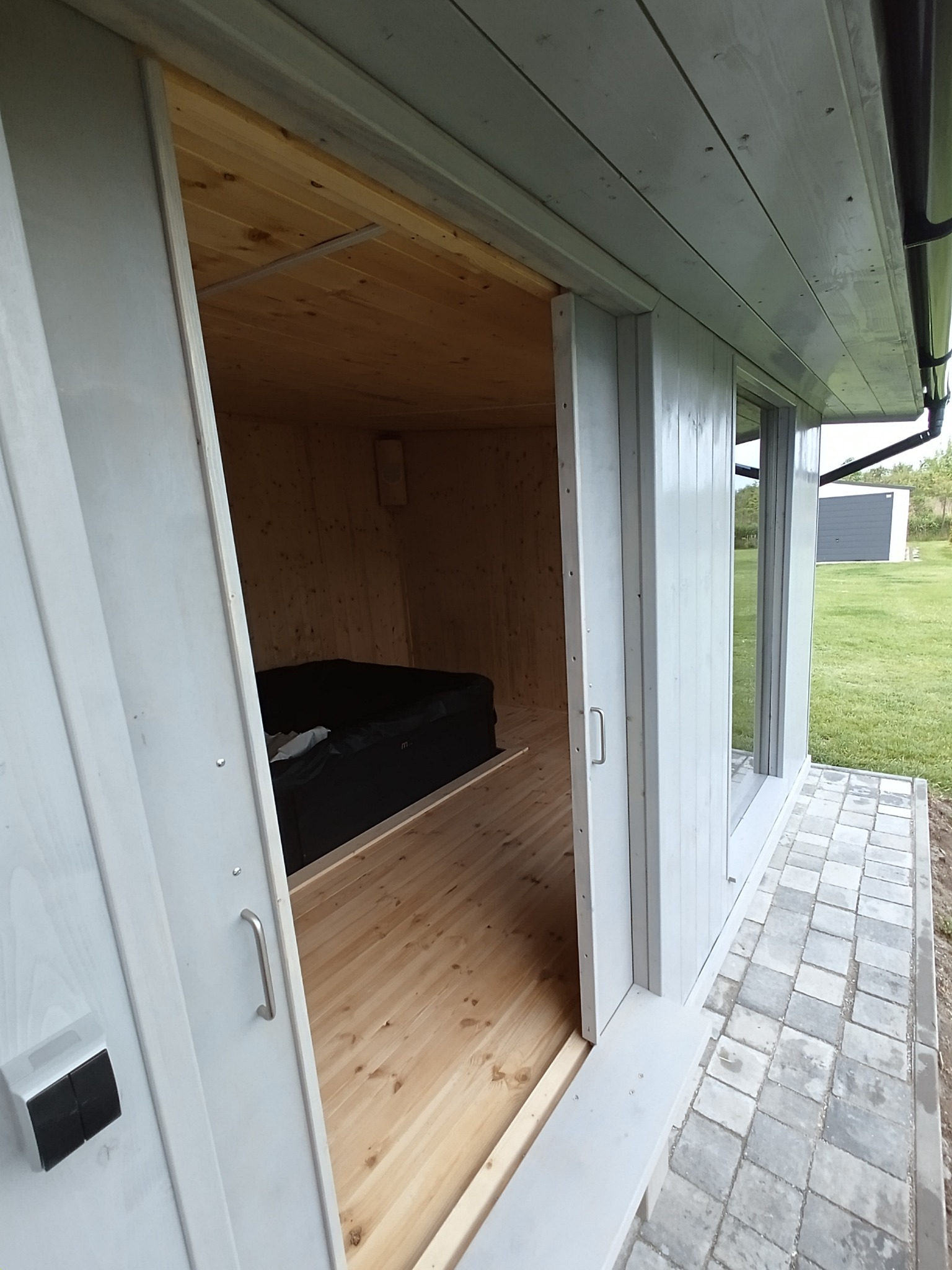 Wnętrze domku spa z jacuzzi, widoczne przez otwarte drzwi. Jasne drewno na ścianach i podłodze, czarna wanna jacuzzi. Zewnętrzna ściana z oknami i kostka brukowa na zewnątrz.