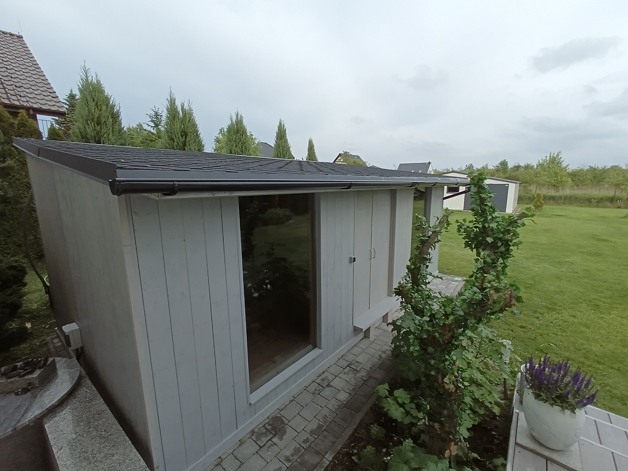 Nowoczesny domek spa z dużym oknem i płaskim dachem, otoczony zielenią. Widoczny fragment tarasu z donicą. Ujęcie z niskiej perspektywy, pochmurne niebo w tle.