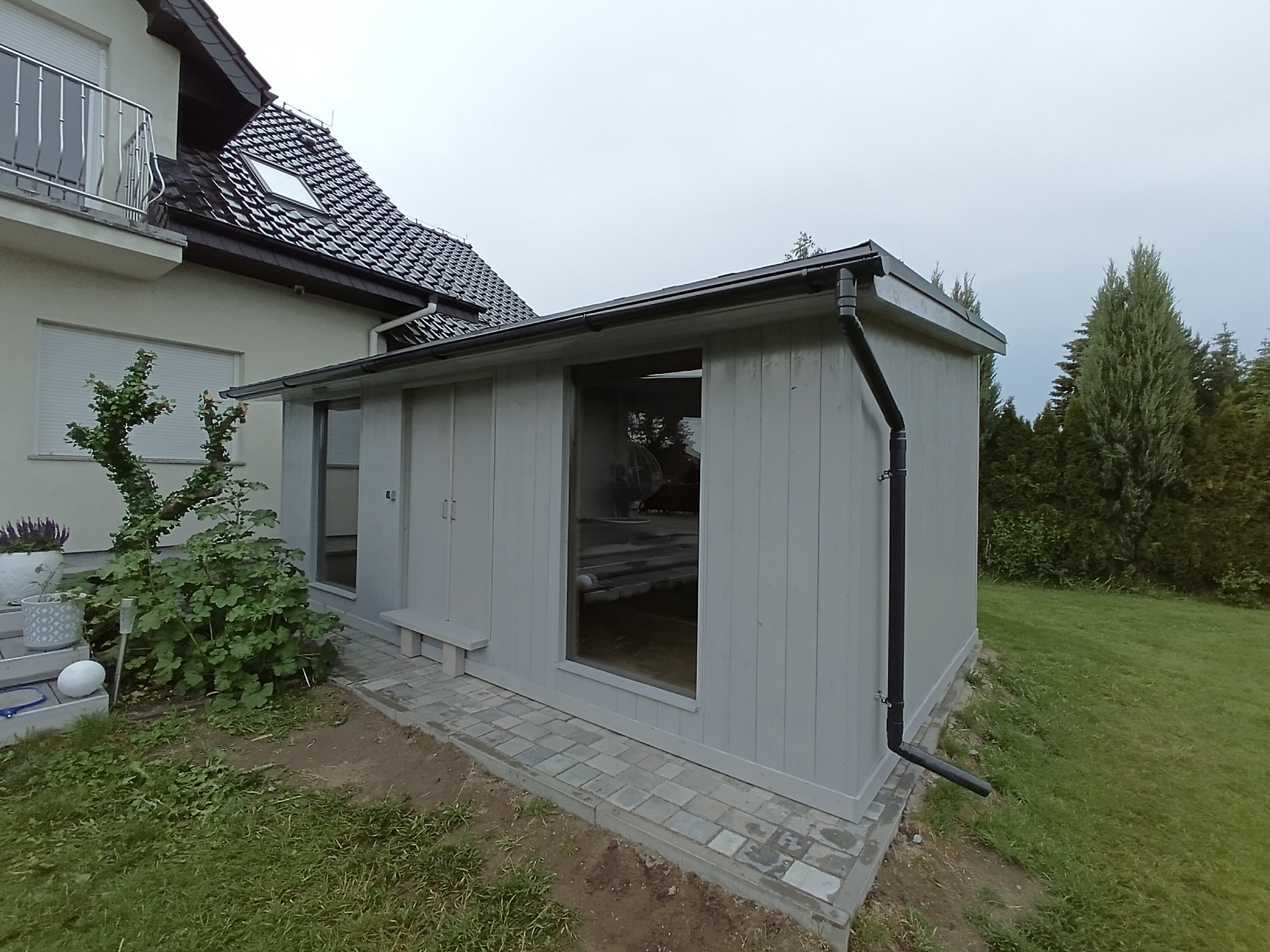 Szary domek spa z sauną i jacuzzi, widoczny z zewnątrz, z oknem ukazującym wnętrze. Obok domu rosną krzewy i trawa, w tle dom z balkonem.