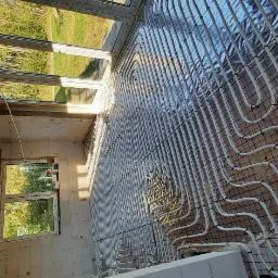 Układanie białych rur ogrzewania podłogowego na folii izolacyjnej z siatką w surowym pomieszczeniu z oknami.