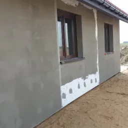 Świeżo nałożony szary tynk na ścianie zewnętrznej domu jednorodzinnego z widocznym ociepleniem przy ziemi i brązowymi oknami, na tle piaszczystego terenu i zieleni.