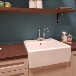 Biały, ceramiczny zlew gospodarczy typu farmhouse z chromowaną baterią w kuchni z beżowymi szafkami i brązowym blatem, dekoracyjne słoiki na półce w tle.