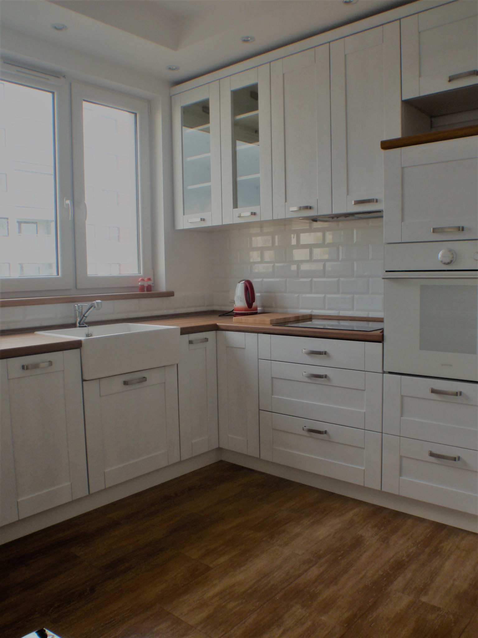 Jasna kuchnia z białymi frontami, drewnianym blatem i ceramicznym zlewem farmerskim, widok na okno z parapetem.