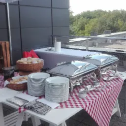 Zewnętrzny bufet z podgrzewaczami, talerzami, koszami z pieczywem, na stole w czerwoną kratę, z grillem w tle na tarasie.