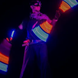 Mężczyzna w masce i stroju scenicznym prezentuje pokaz świetlny z użyciem rekwizytów LED w kolorach niebieskim, pomarańczowym i zielonym na ciemnym tle.