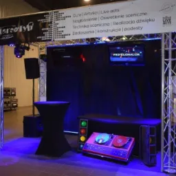 Scena z nagłośnieniem i oświetleniem, widoczne elementy konstrukcyjne, baner reklamowy z napisem StereoTyp, gramofon, kolumny, stolik koktajlowy, monitory i oświetlenie sceniczne w kolorze niebieskim.