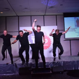 Czterech mężczyzn w czarnych strojach skacze synchronicznie na scenie podczas wydarzenia firmowego, z logo firmy 'We are Tupp' w tle i wyświetlanymi slajdami na temat ekologii.