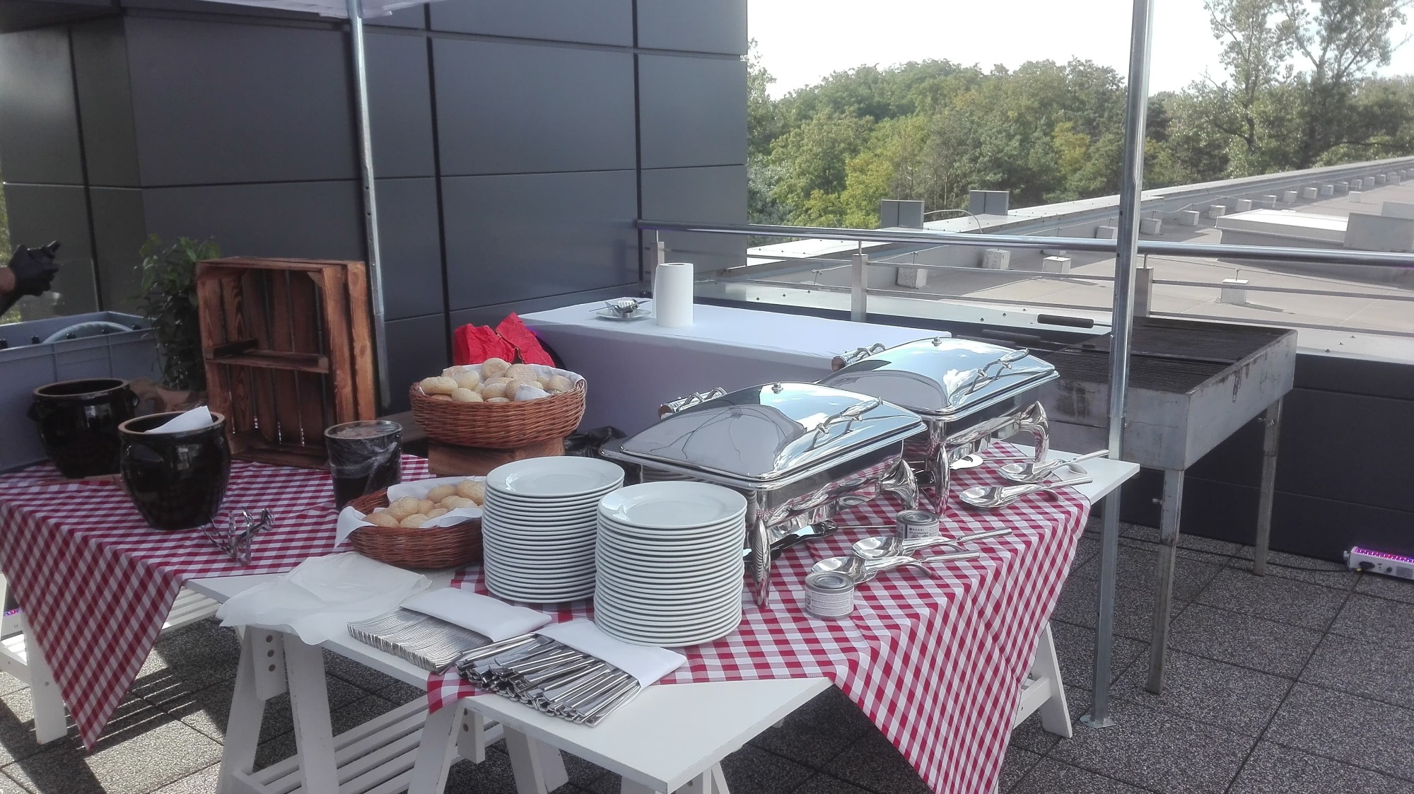 Zewnętrzny bufet z podgrzewaczami, talerzami, koszami z pieczywem, na stole w czerwoną kratę, z grillem w tle na tarasie.
