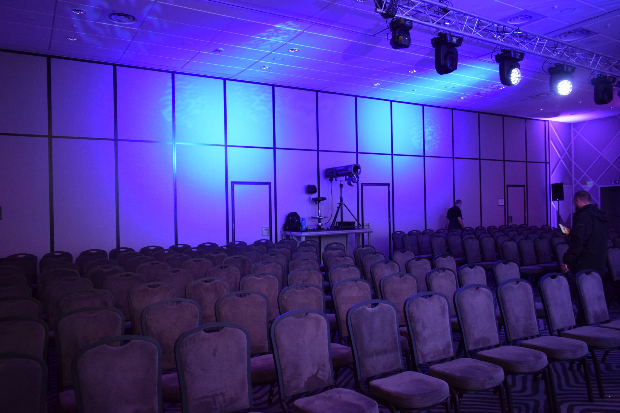 Sala konferencyjna przygotowana do wydarzenia, oświetlona niebieskim światłem, z rzędami krzeseł i sprzętem nagłośnieniowym na scenie.