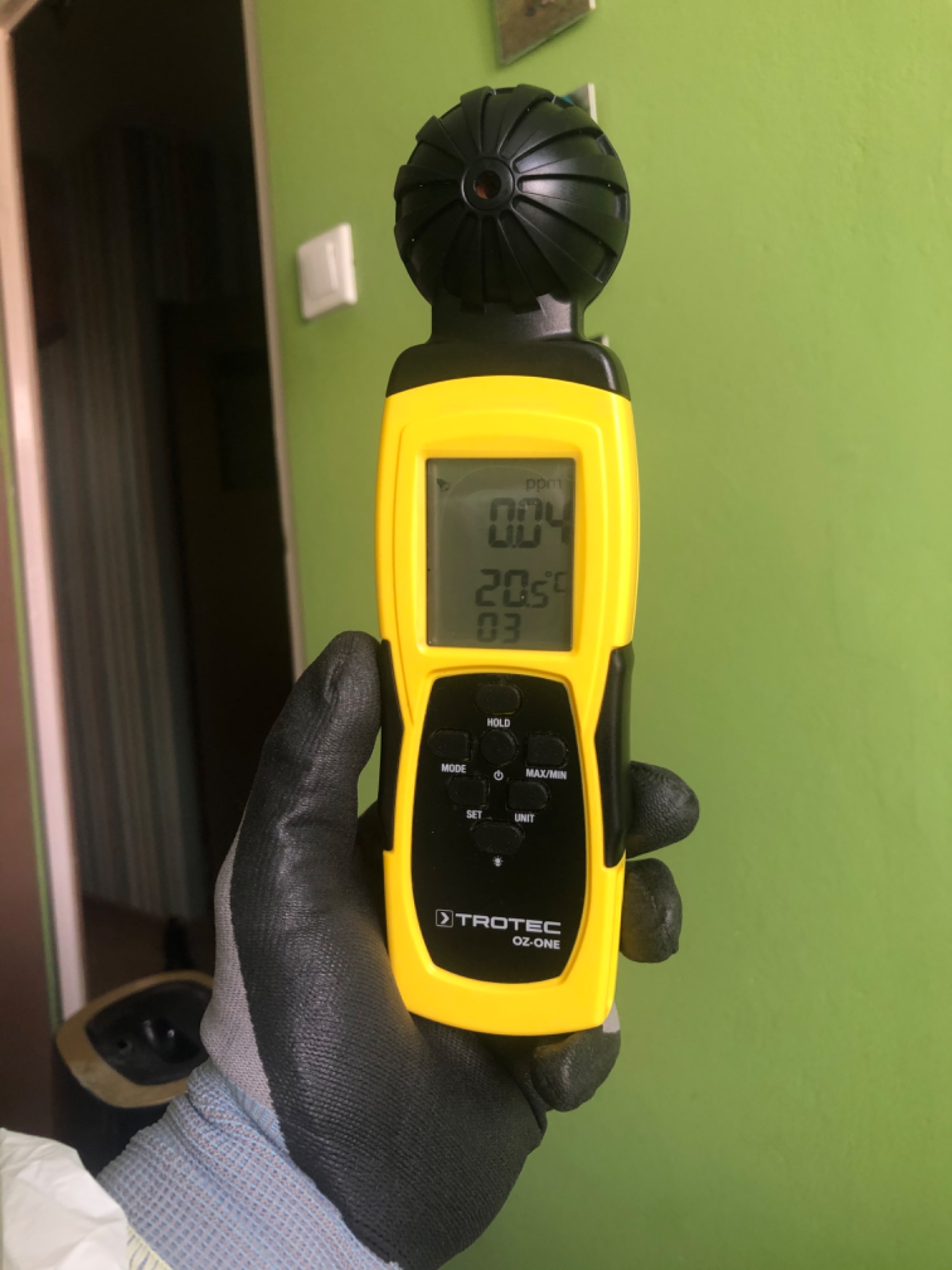 Miernik ozonu Trotec OZ-ONE w dłoni w rękawicy ochronnej, wyświetlający odczyt 0.04 ppm na tle zielonej ściany i otwartego przejścia.