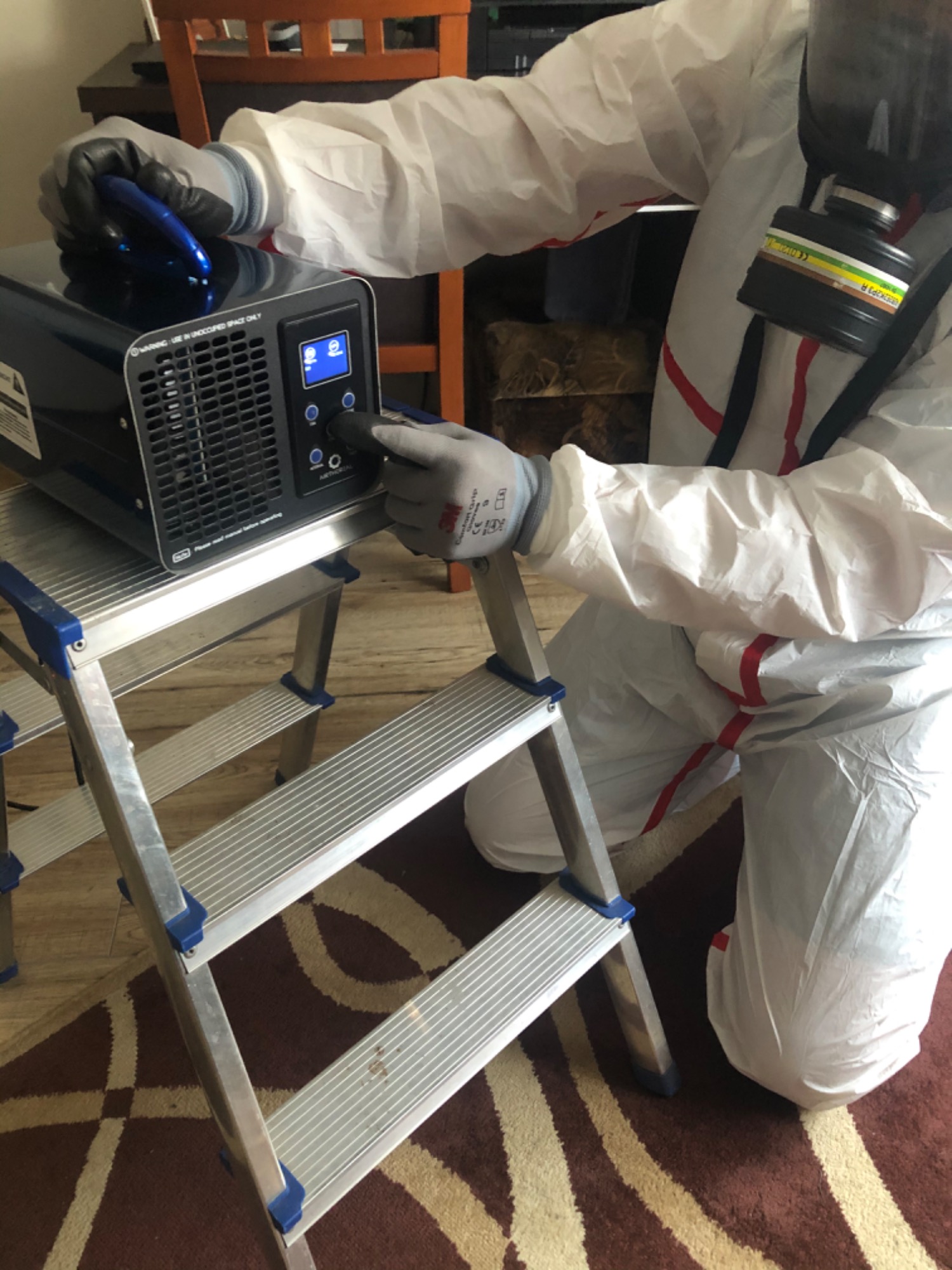 Osoba w kombinezonie ochronnym i masce obsługuje ozonator na aluminiowej drabinie w pomieszczeniu.