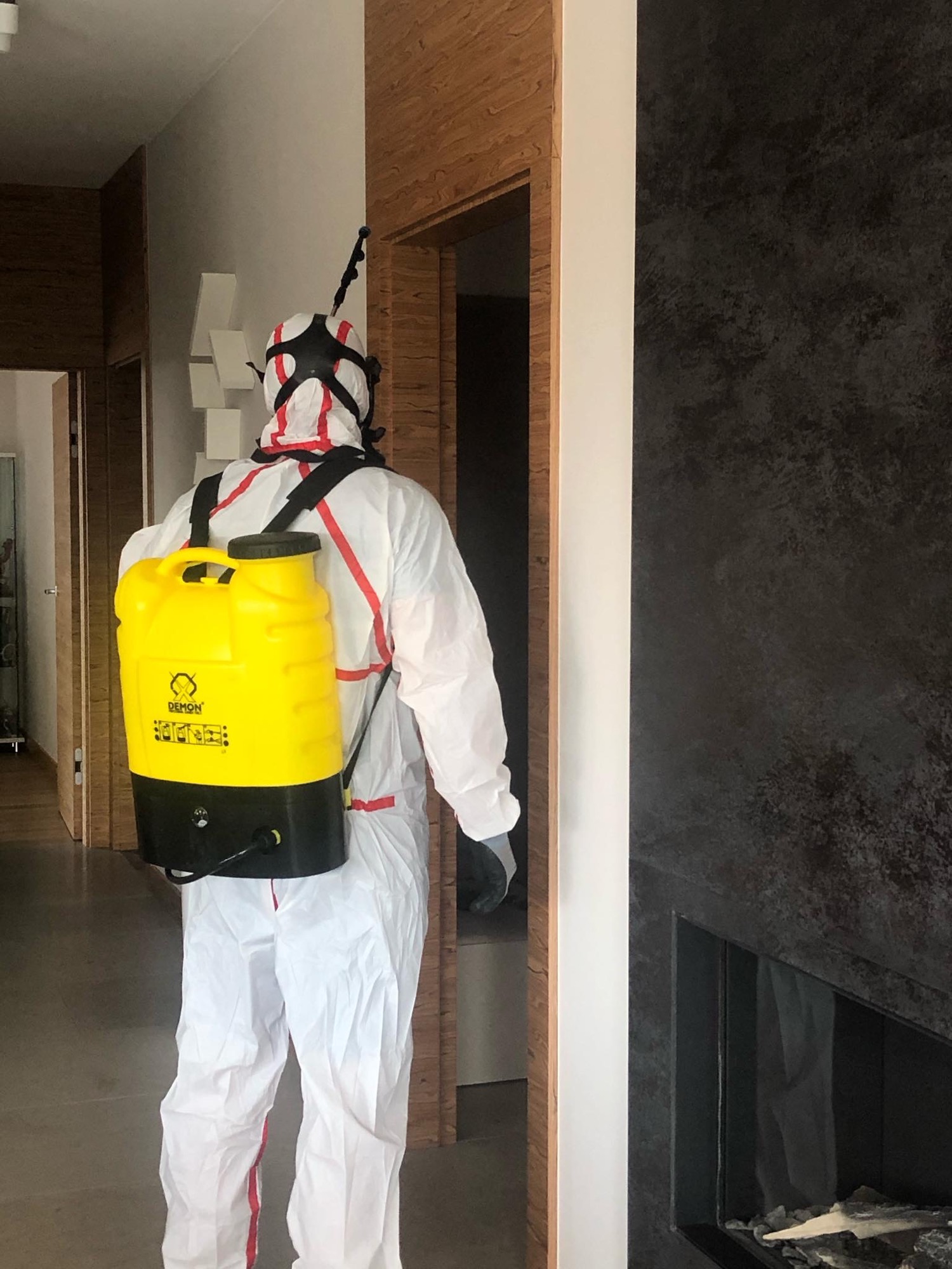 Osoba w kombinezonie ochronnym i masce z żółtym opryskiwaczem na plecach, przygotowująca się do dezynfekcji wnętrza z ciemnym kominkiem.