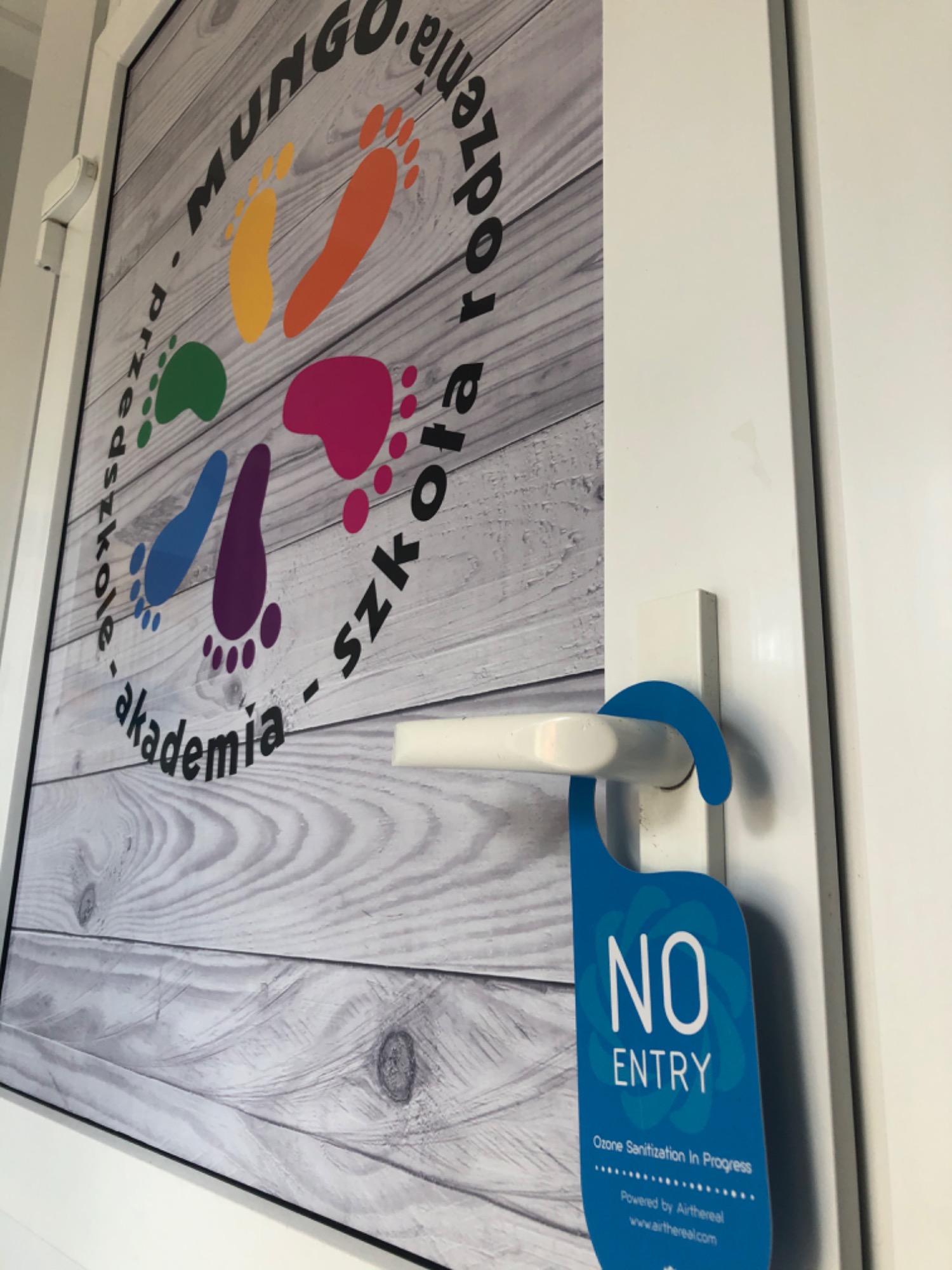 Tabliczka na drzwiach z napisem 'NO ENTRY, Ozone Sanitization In Progress' wisząca na klamce, obok plakatu przedszkola 'Akademia Mungo - szkoła rodzenia' z kolorowymi odciskami stóp na tle...