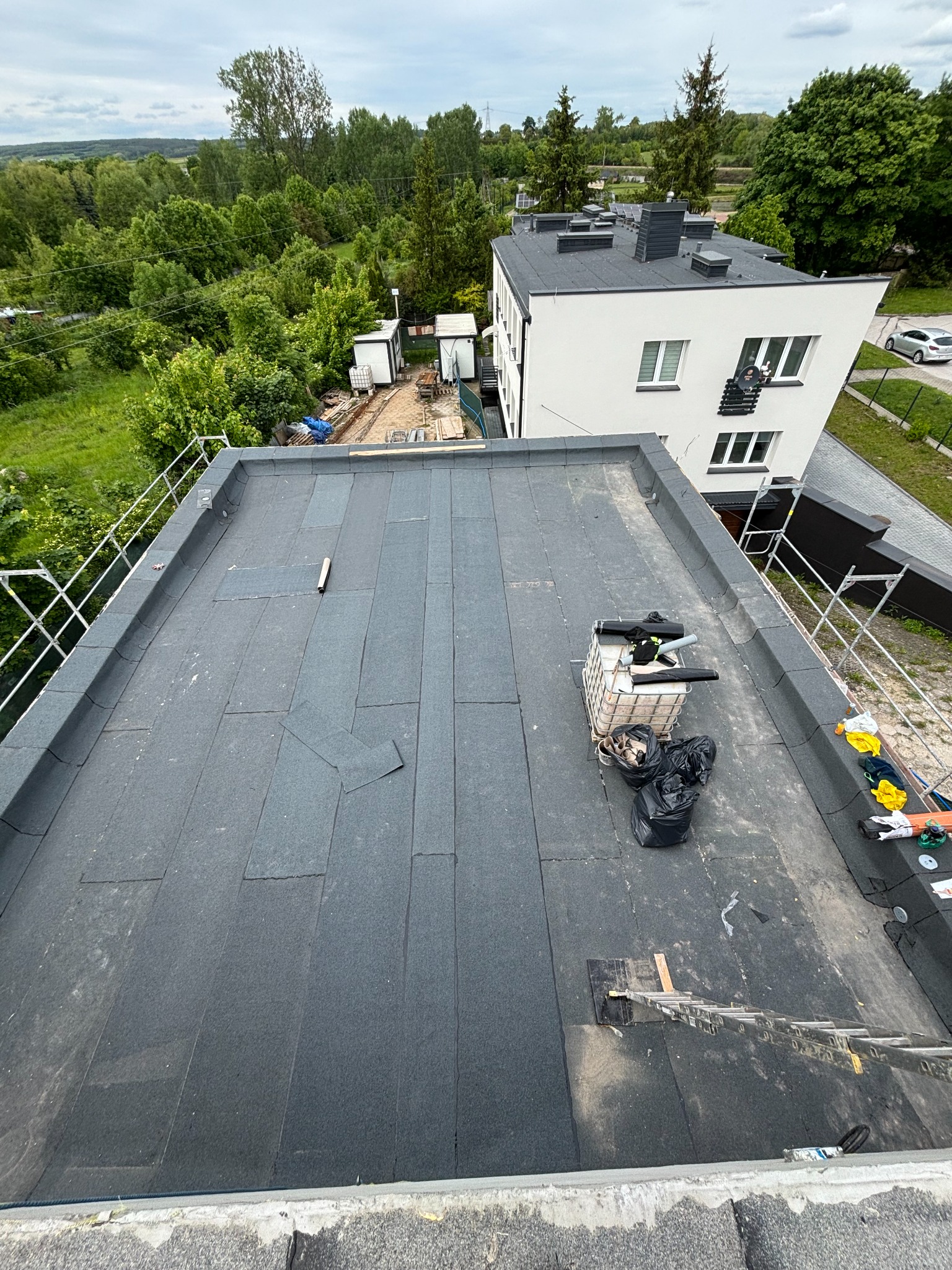Czarny, płaski dach pokryty papą termozgrzewalną, z widocznymi akcesoriami dekarskimi i fragmentem budynku w tle. Ujęcie z góry, w dzień.