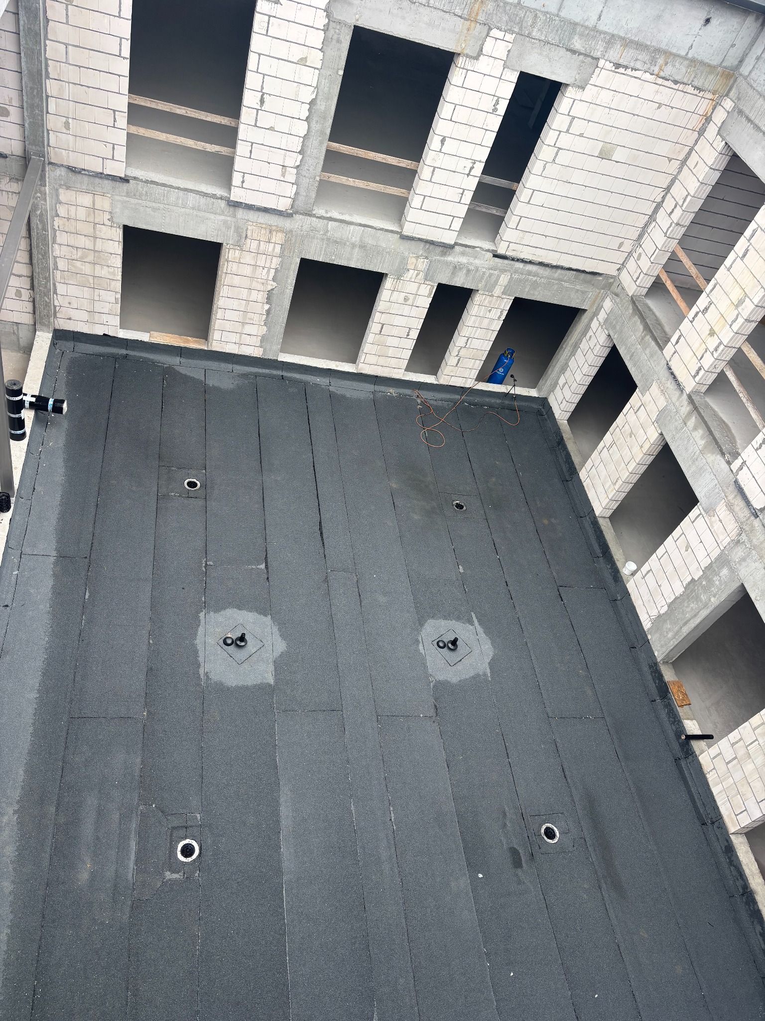 Czarny, płaski dach pokryty papą termozgrzewalną na surowym budynku z betonu komórkowego. Widoczne wpusty dachowe i butla z gazem.