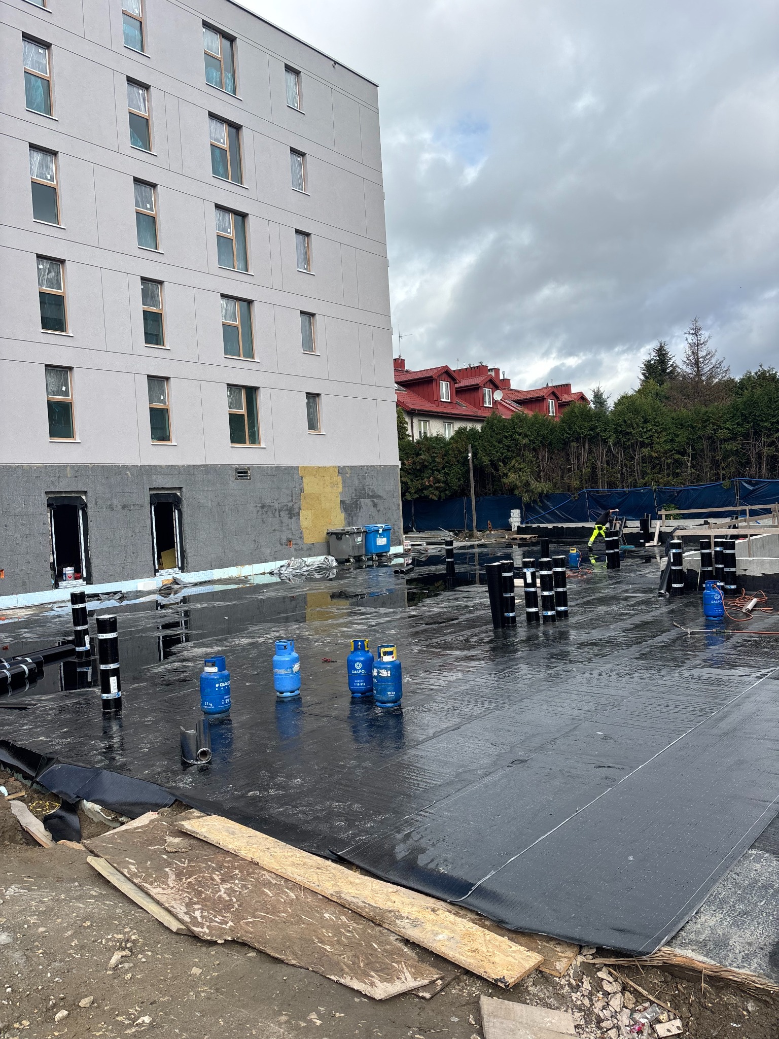 Czarna papa na dachu budynku wielorodzinnego w trakcie prac. Widoczne butle gazowe, zrolowana papa i pracownik w oddali. Pochmurne niebo w tle.