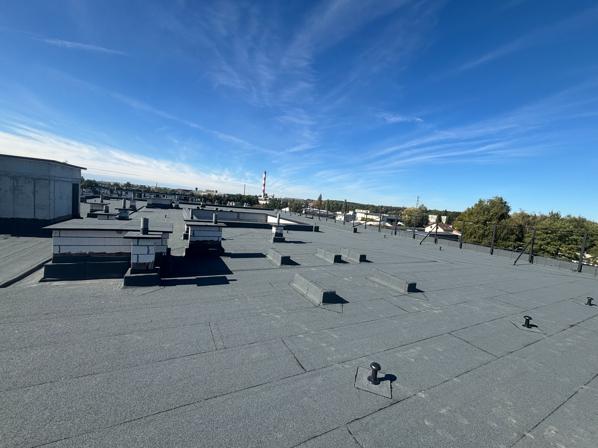 Płaski dach pokryty papą termozgrzewalną w odcieniach szarości, z widocznymi kominami i elementami wentylacyjnymi. W tle błękitne niebo z delikatnymi chmurami i zabudowa miejska.
