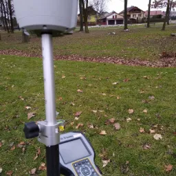 Geodezyjny odbiornik GNSS Trimble na statywie, z kontrolerem w tle trawnik z opadłymi liśćmi i zabudowa jednorodzinna.