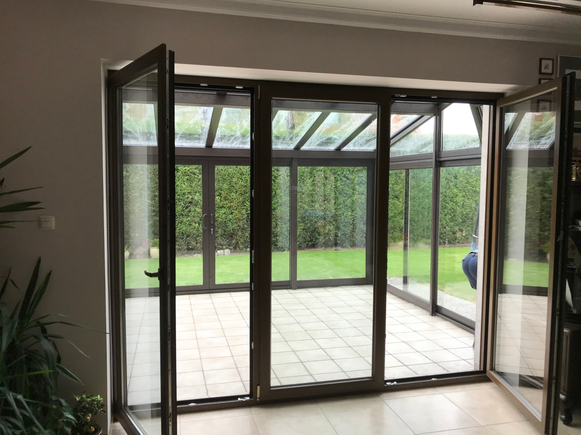 Zadaszony taras z przesuwnymi drzwiami szklanymi z brązowymi ramami, widok na zielony trawnik i żywopłot, częściowo otwarte drzwi po lewej i prawej stronie.