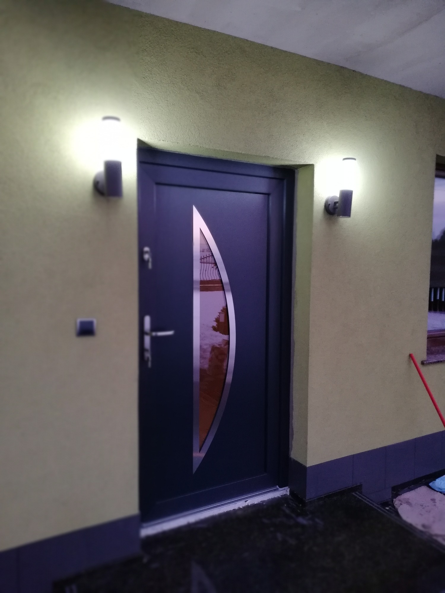 Grafitowe drzwi wejściowe z owalnym przeszkleniem, oświetlone dwoma kinkietami na teksturowanej, oliwkowej ścianie domu.