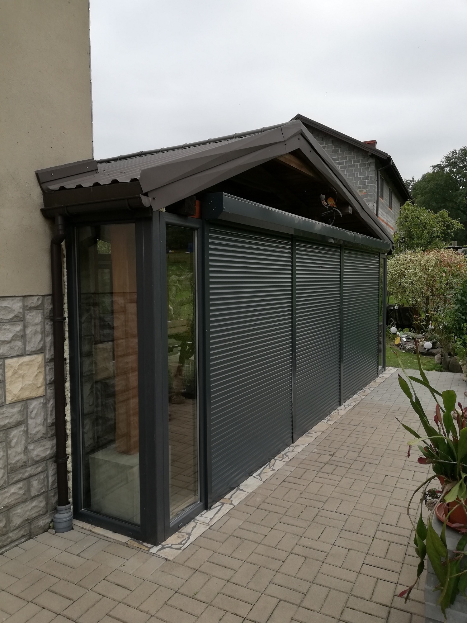 Zadaszony taras z ciemnymi roletami zewnętrznymi i przeszklonymi elementami, widok z zewnątrz na tle kostki brukowej i roślinności ogrodowej.