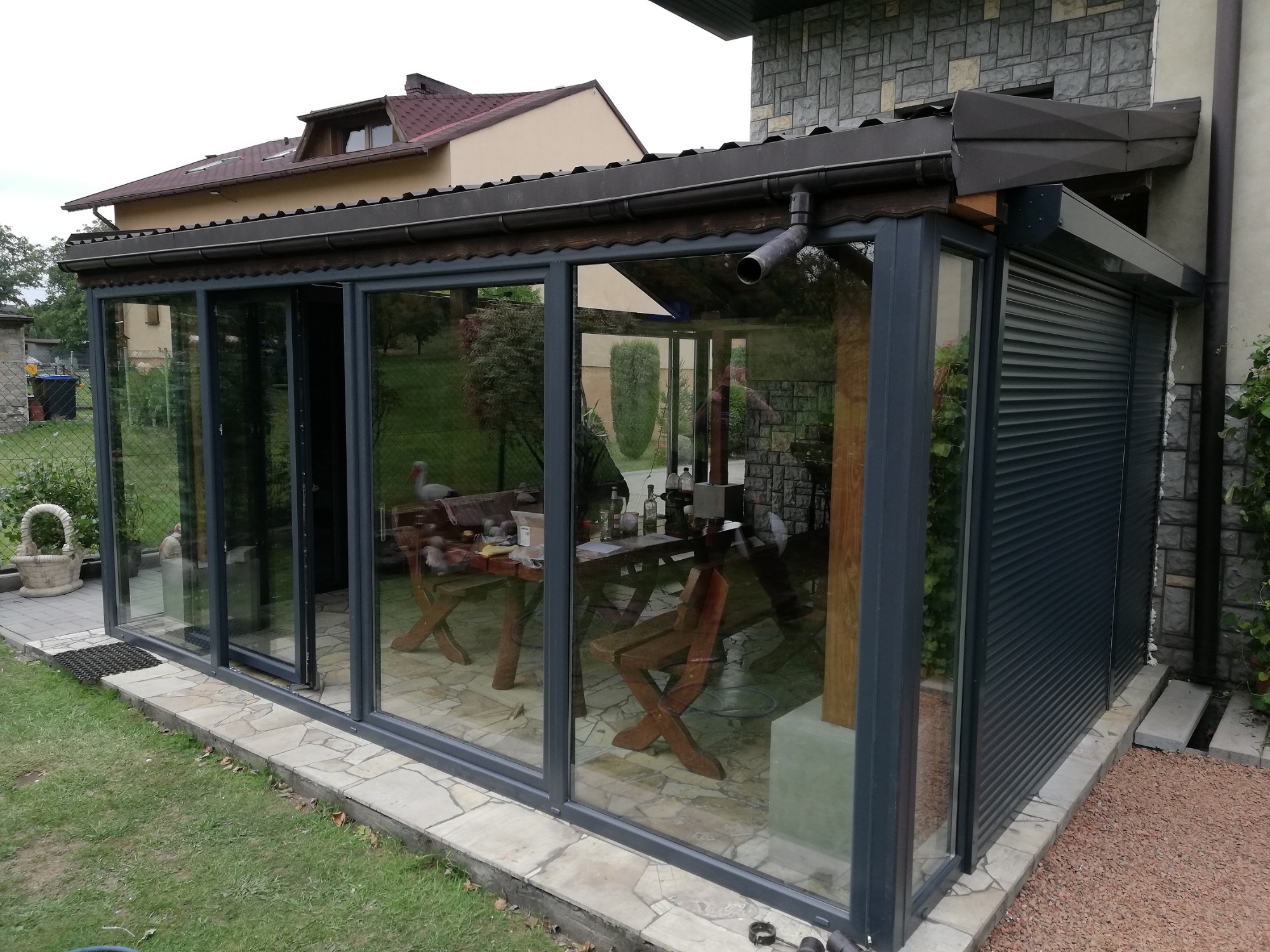 Zadaszony taras z przesuwnymi szklanymi ścianami w szarej ramie i częściowo zasłoniętymi zewnętrznymi roletami, widoczny drewniany stół i krzesła wewnątrz.